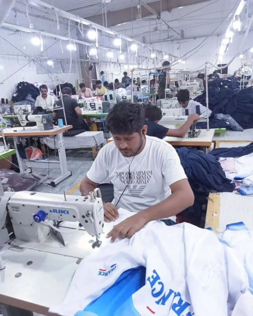 Garment factory workers operating industrial sewing machines during bulk sportswear assembly at Feel It Creation manufacturing unit.