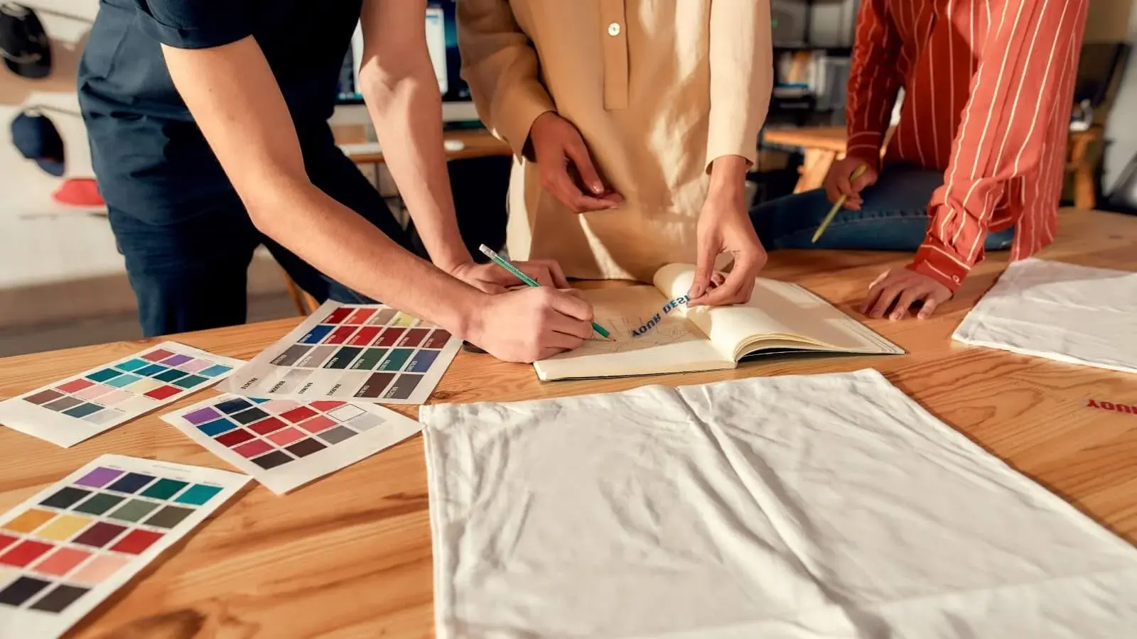 Fashion designers collaborating at a design table with pantone fabric swatches, tech pack sketches, and apparel samples at Feel It Creation.