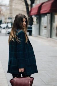 Woman walking down a city street wearing an oversized dark green plaid wool coat and carrying a burgundy studded handbag manufactured by Feel It Creation.